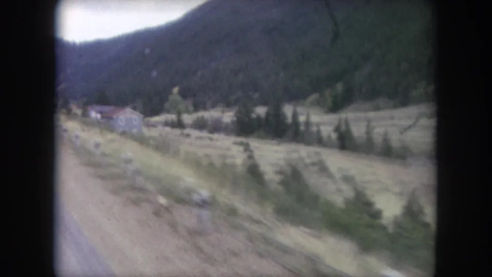 1962 Winter Park, Colorado home-movie frame showing a passing roadside valley with pine-covered hills and small distant structures.