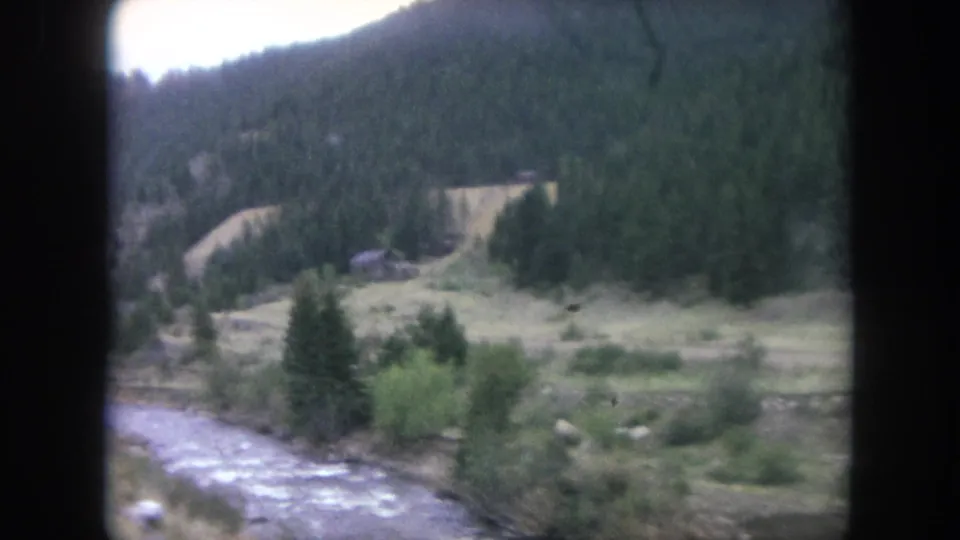 1962 Winter Park, Colorado frame of a river running through a wooded mountain valley, seen through dark vehicle-window borders.