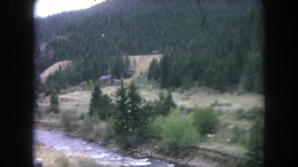 1962 Winter Park, Colorado frame showing riverbank shrubs, evergreens, and a small structure within a mountain valley landscape.