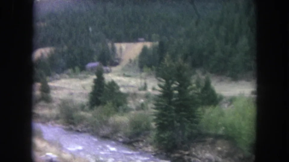 1962 Winter Park roadside view with creek water, evergreens, meadow, and a small structure near the forest edge.