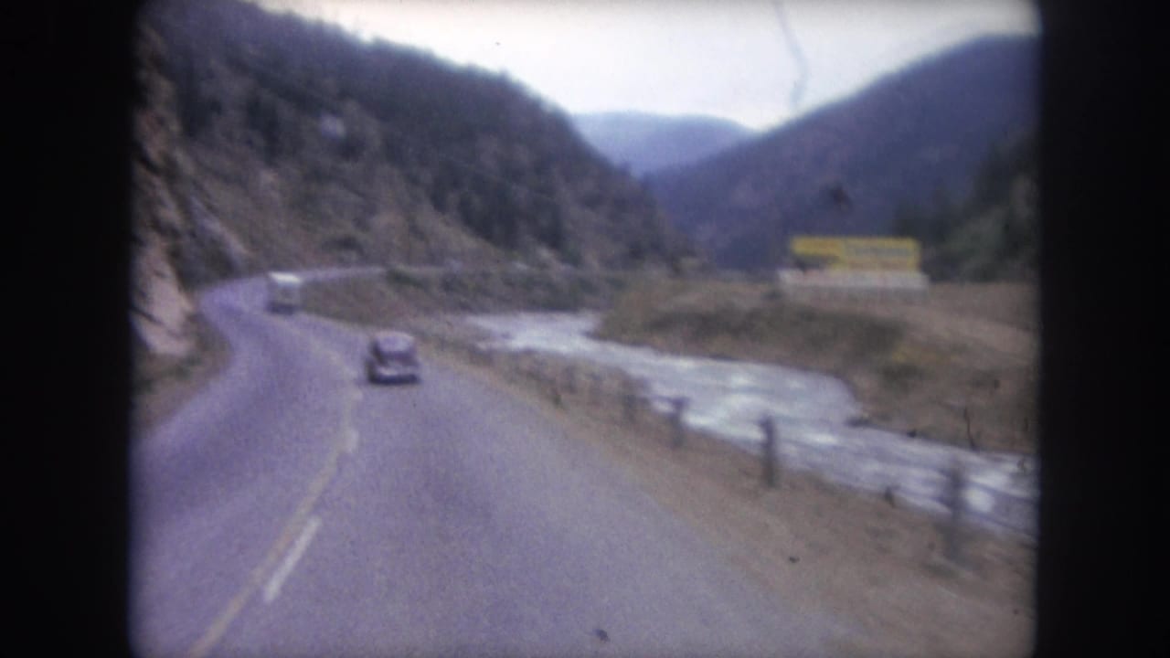 Two-lane mountain highway beside a river with a dark car ahead and forested slopes.