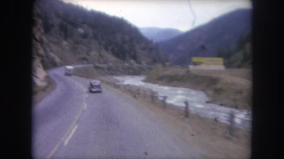 1962 8mm view of a two-lane highway near Winter Park, Colorado, with a dark sedan, river, rocky hillside, and distant yellow sign.