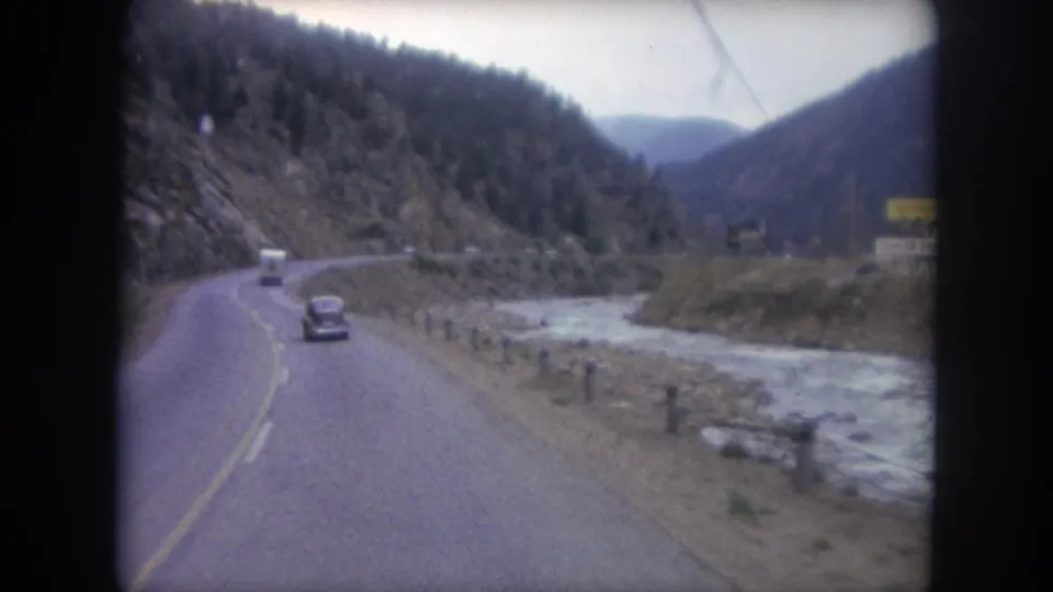 A 1962 home-movie frame follows traffic on a two-lane mountain road near Winter Park, with riverbank posts and wooded hills visible.
