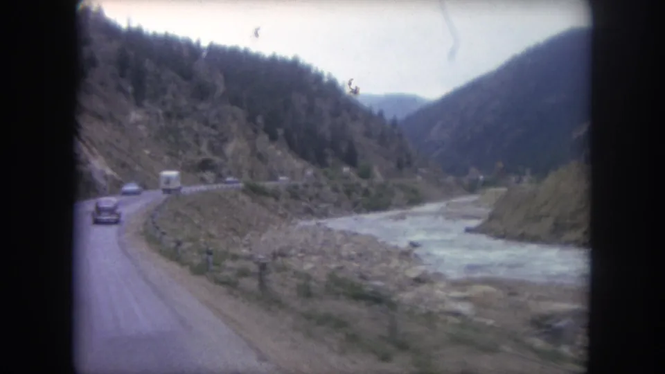 Home-movie view from a moving car on a 1962 Winter Park road, looking toward vehicles, a rocky riverbed, and wooded hills.