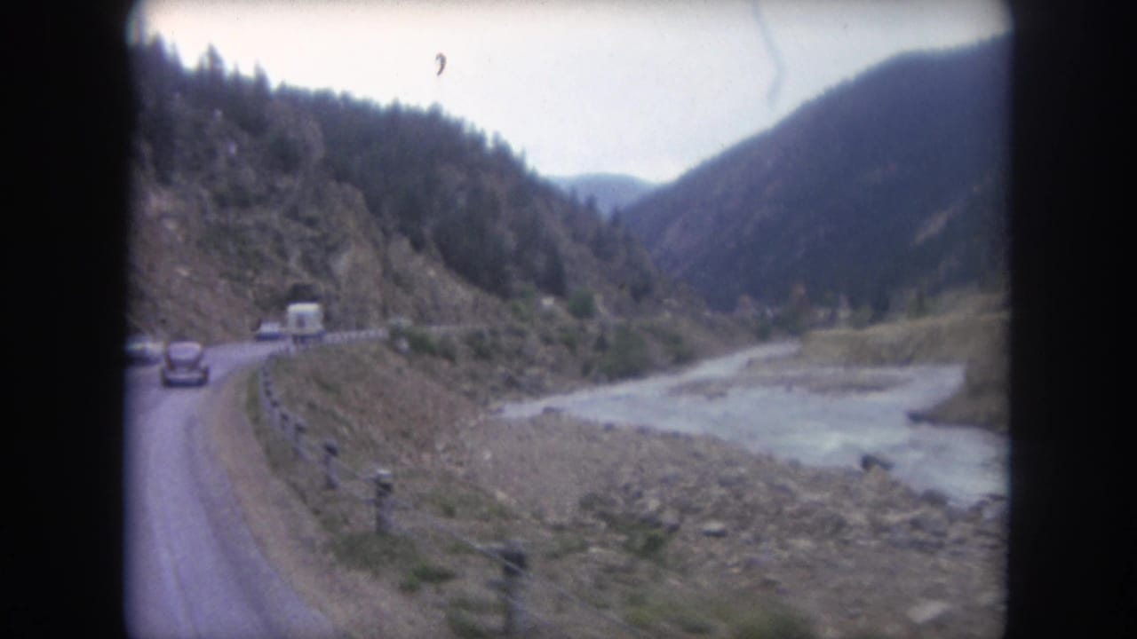 1962 Colorado 8mm frame showing a mountain road curving beside a river, with cars, a white trailer, guard posts, and forested slopes.