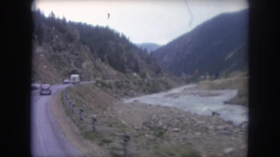 1962 Colorado 8mm frame showing a mountain road curving beside a river, with cars, a white trailer, guard posts, and forested slopes.