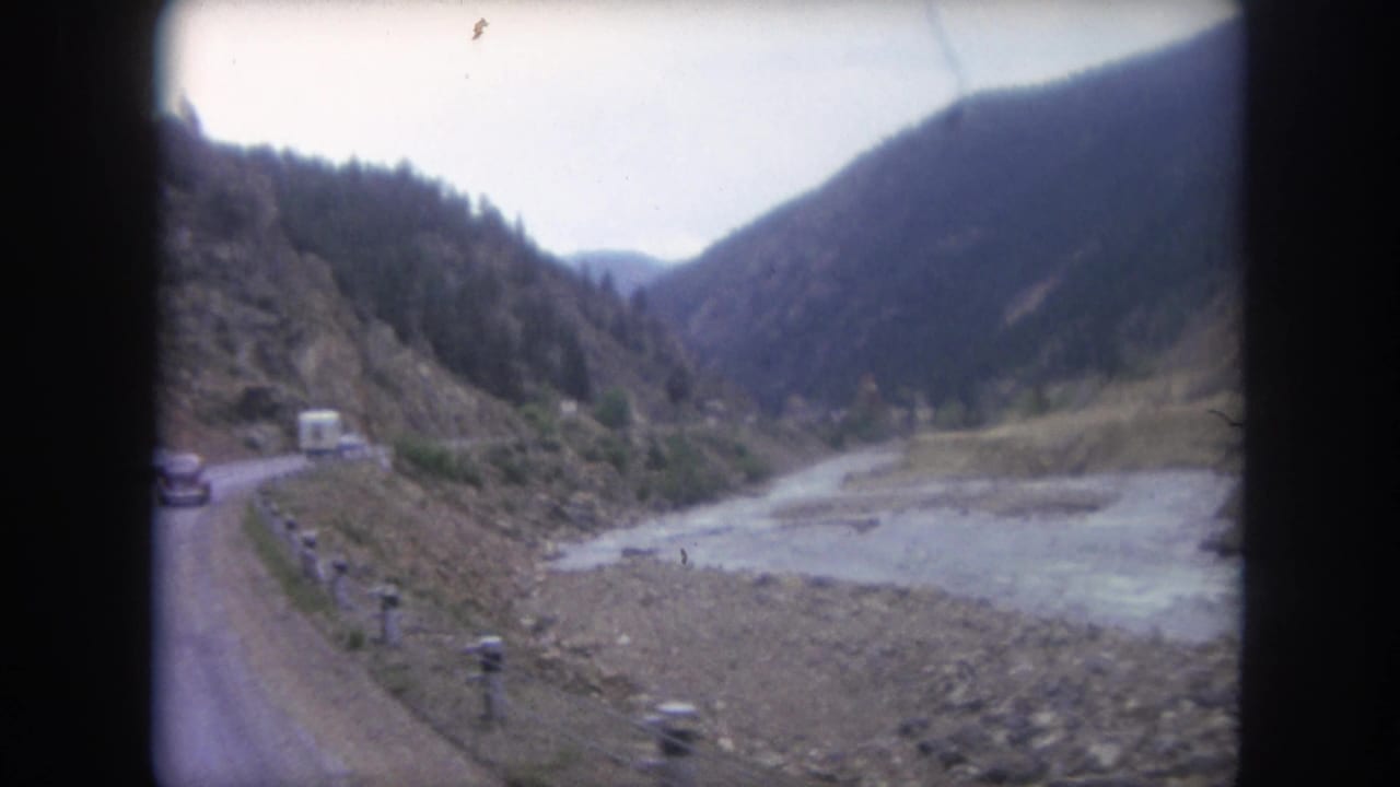 1962 Colorado 8mm frame of vehicles on a two-lane mountain road beside a pale river and steep forested valley walls.