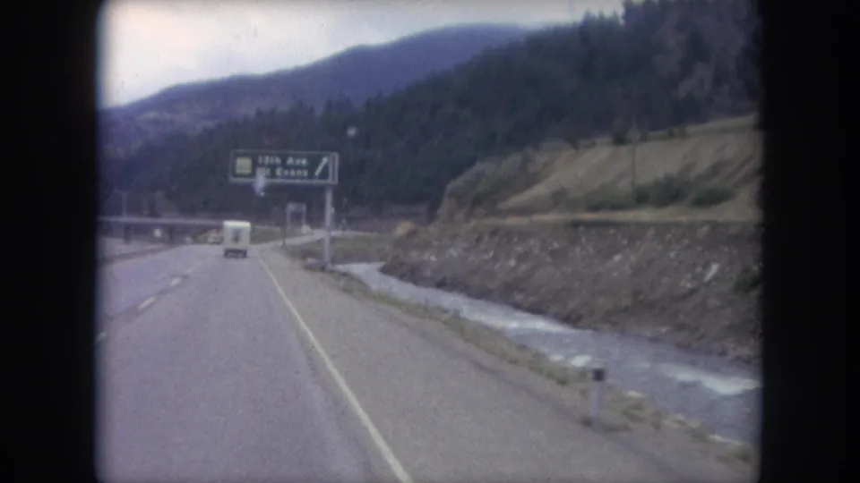 1962 Colorado 8mm frame showing a vehicle ahead on a mountain road approaching an overhead sign for 13th Ave and Mt. Evans.