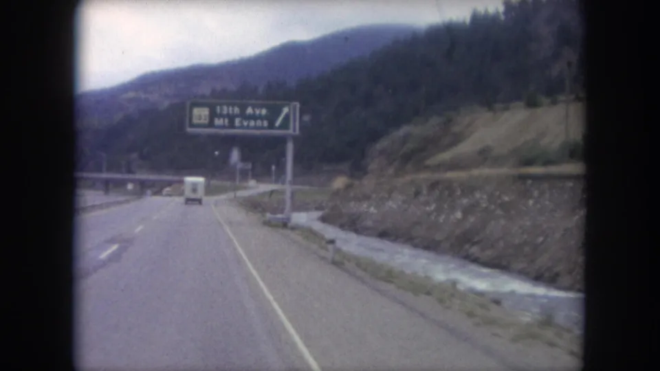 1962 Colorado 8mm frame with an overhead 13th Ave and Mt. Evans road sign, a white vehicle ahead, and a stream beside the highway.