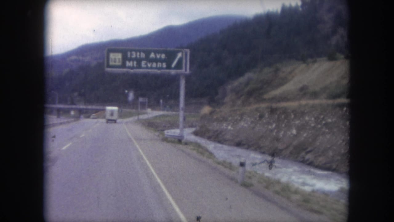 Mountain highway seen from a moving vehicle with a green road sign and white van ahead.