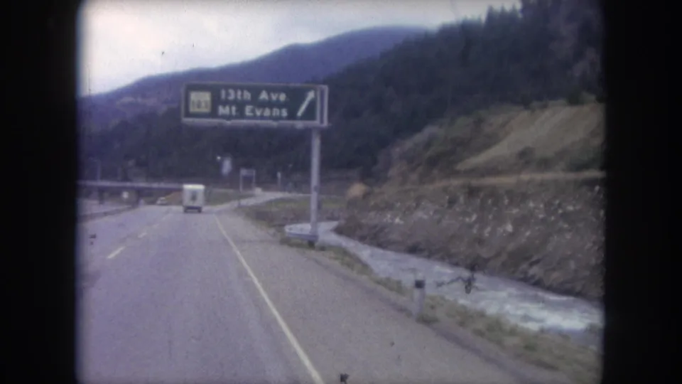 1962 8mm view of a Colorado mountain highway with a Route 103 sign for 13th Ave and Mt. Evans, forested slopes, and a white van ahead.