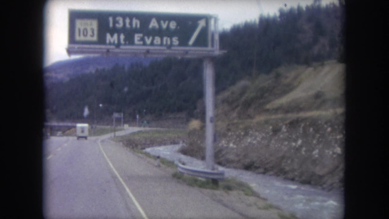 1962 Colorado road footage passes beneath a green Route 103 guide sign for 13th Ave and Mt. Evans, with a stream and wooded slope beside the road.