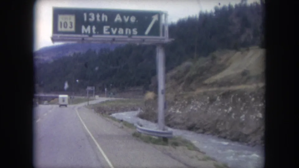 1962 Colorado road footage passes beneath a green Route 103 guide sign for 13th Ave and Mt. Evans, with a stream and wooded slope beside the road.