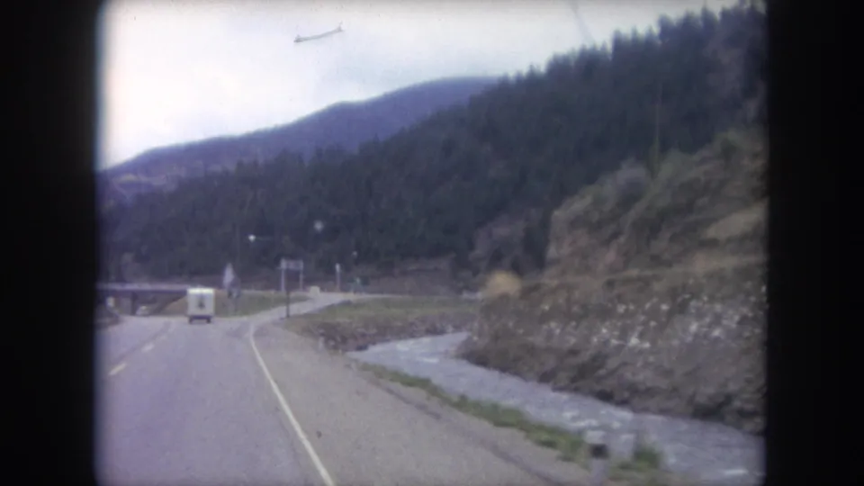 1962 8mm driving view along a Colorado mountain highway after passing the road sign, with a white van ahead and a stream along the shoulder.