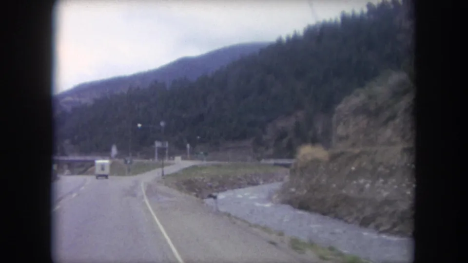 1962 Colorado mountain-road footage shows the highway opening ahead, a white van near the center, and a stream running beside the rocky shoulder.