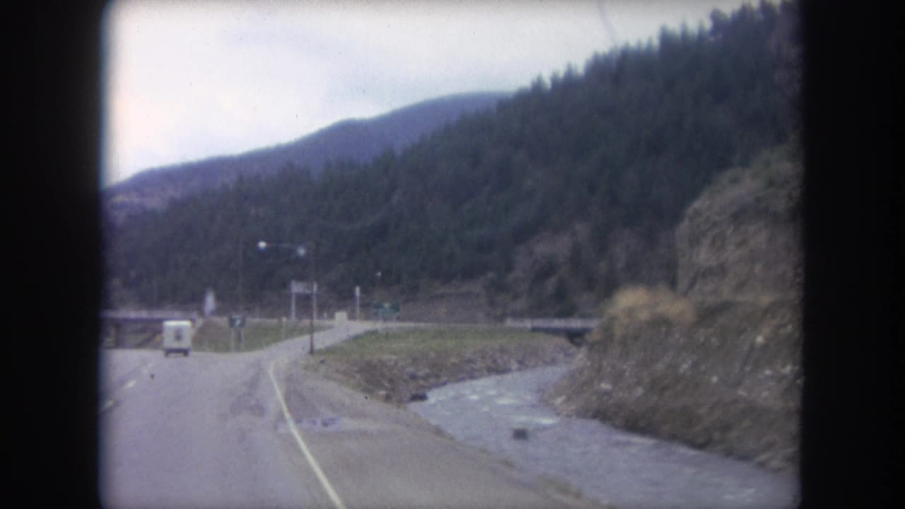 Curving mountain road beside a rocky stream with forested hills and a white trailer ahead.