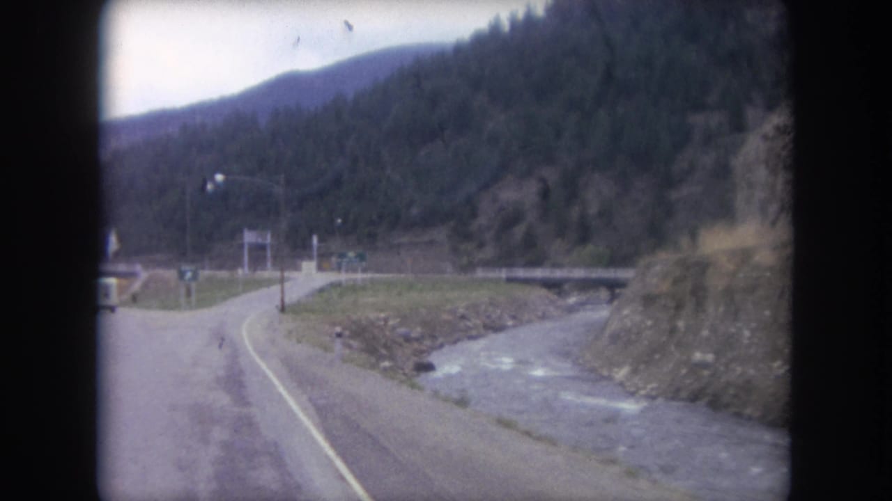 Forward-facing 8mm car view near Winter Park, Colorado, with a road ramp or fork, creek, bridge, signs, and dark evergreen slopes.