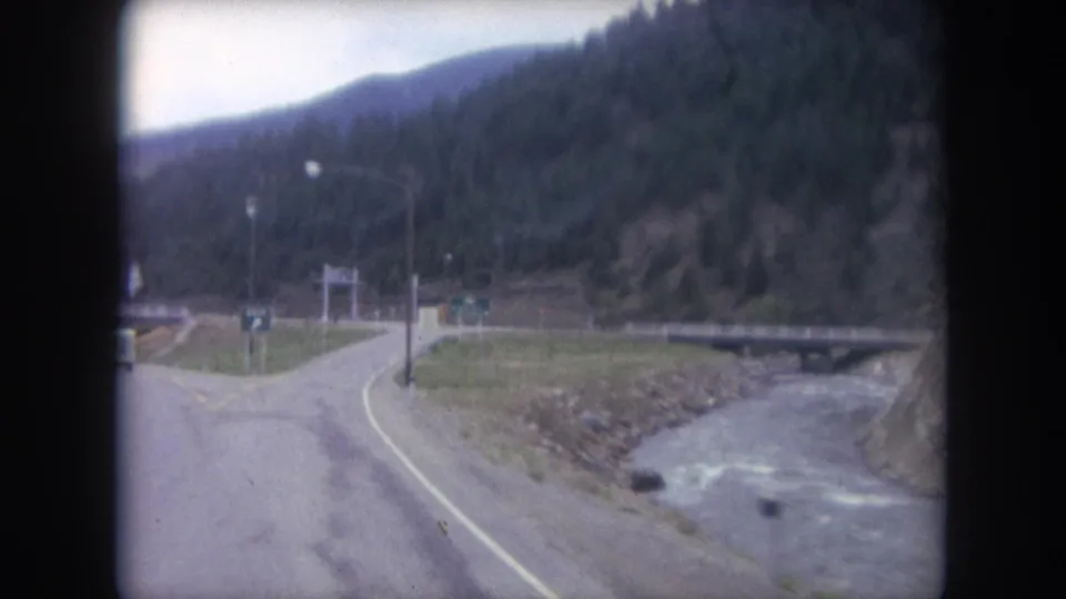 1962 home-movie frame from a moving car in Winter Park, Colorado, showing a curving road, creek, bridge approach, utility pole, and wooded hillside.