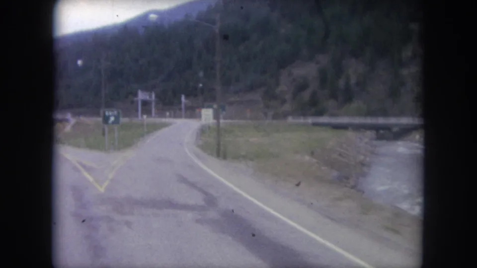 Final 1962 Winter Park road-travel frame showing a ramp-like lane, creek to the right, bridge, roadside signs, and forested mountain terrain.