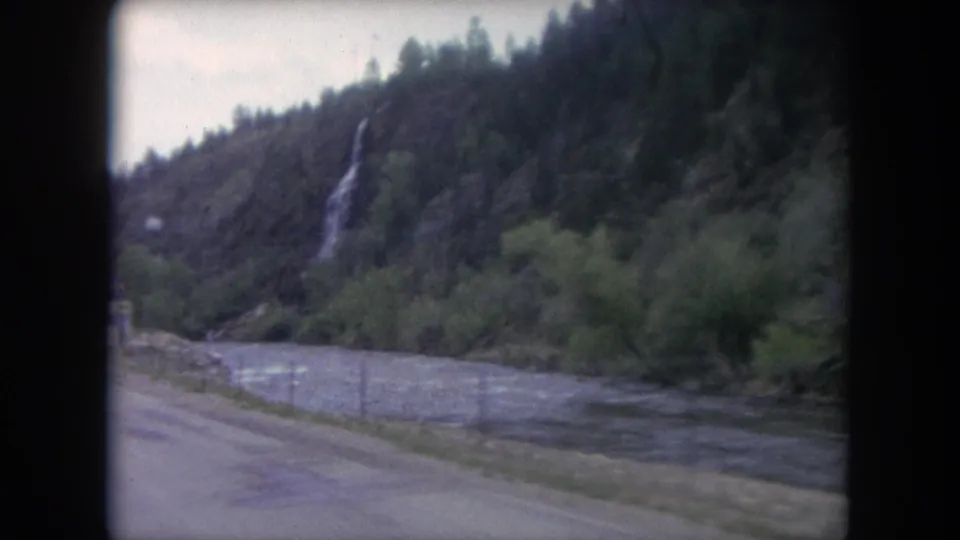 Winter Park, Colorado mountain scenery from 1962 showing a roadside river, rocky slope, trees, and a narrow waterfall in soft 8mm focus.