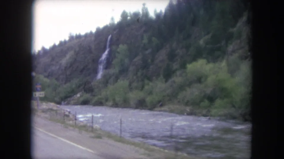 A 1962 Winter Park, Colorado roadside view of a waterfall dropping through a rocky, wooded slope above a rushing river.