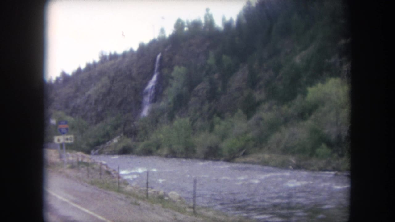 1962 8mm view near Winter Park, Colorado, showing a roadside river, wooded mountain slope, and distant waterfall from a moving vehicle.