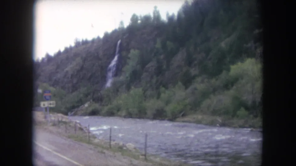 1962 8mm view near Winter Park, Colorado, showing a roadside river, wooded mountain slope, and distant waterfall from a moving vehicle.