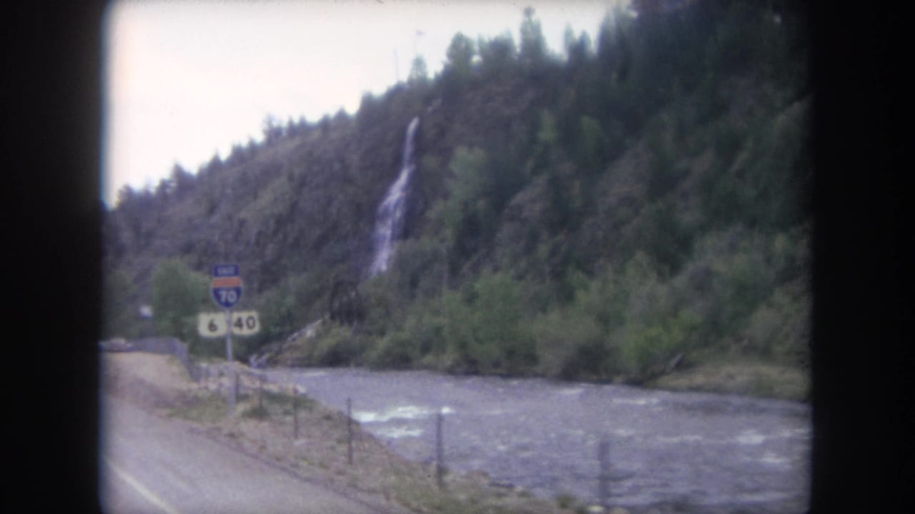 Moving-car view of a Colorado mountain river with a waterfall and U.S. Route 6 and 40 signage near an Interstate 70 East marker.