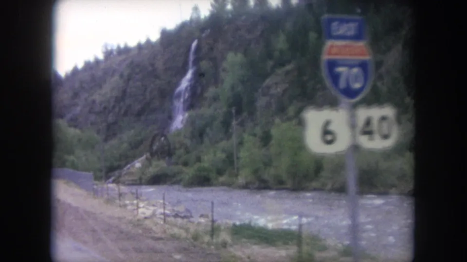 Close moving 8mm frame of U.S. Route 6 and 40 markers and an Interstate 70 East sign, with a Colorado mountain river and waterfall behind.