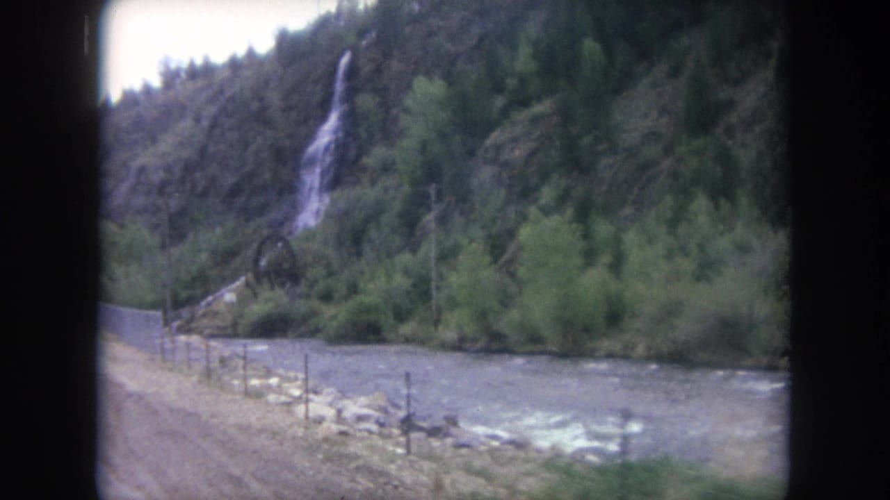 Roadside river with a small waterfall descending a wooded rocky hillside.