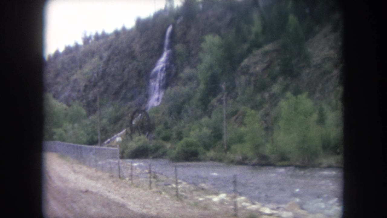 A Winter Park, Colorado roadside scene shows a narrow waterfall, green vegetation, a river, fencing, and utility poles from a moving 8mm view.