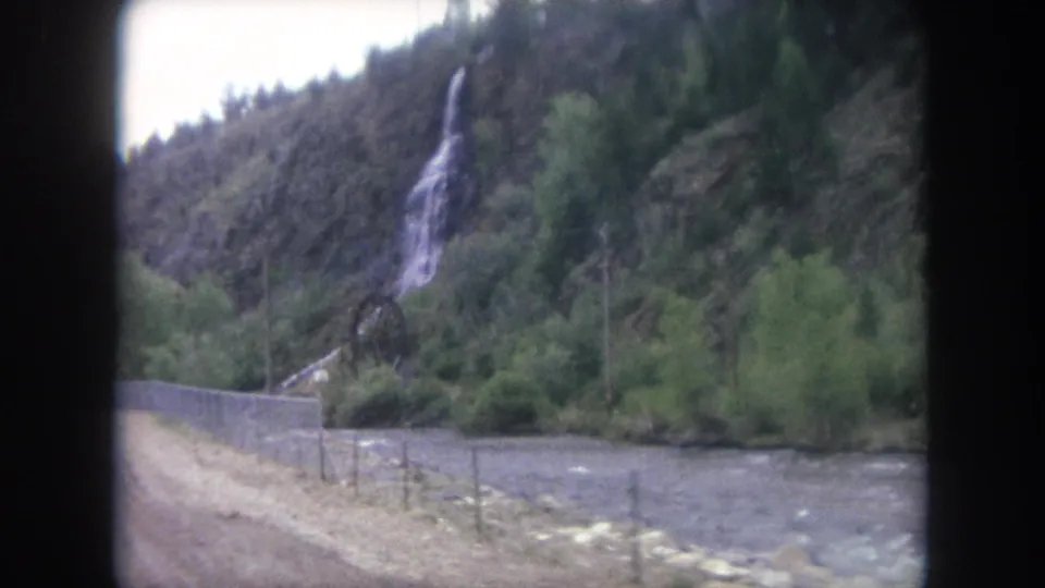 A Winter Park, Colorado roadside scene shows a narrow waterfall, green vegetation, a river, fencing, and utility poles from a moving 8mm view.