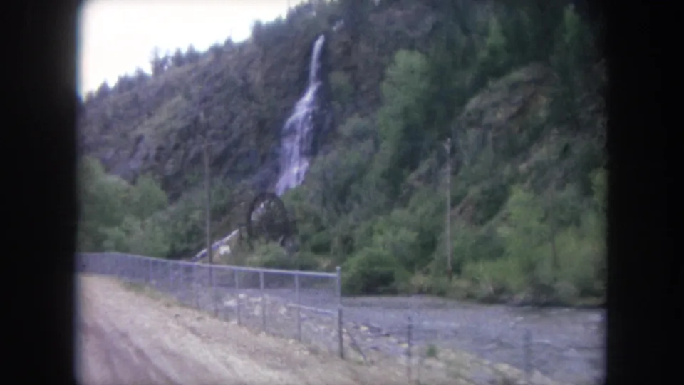 The 1962 home-movie frame shows the vehicle nearing a fenced roadside section below a rocky hillside waterfall in Winter Park, Colorado.