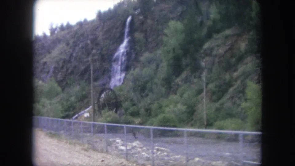 A final 8mm frame from Winter Park, Colorado shows a river behind roadside fencing, utility poles, trees, and a thin waterfall on the hill.