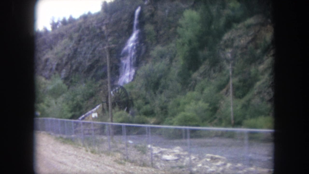 1962 8mm view in Winter Park, Colorado, showing a hillside waterfall above trees, fencing, utility poles, and a small structure.