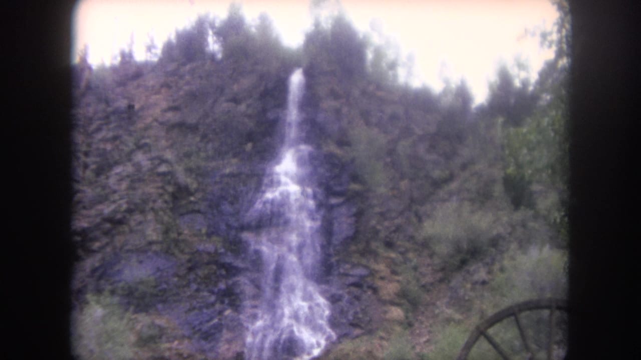 Closer view of a narrow waterfall dropping down a rocky slope with trees and part of a waterwheel at right.