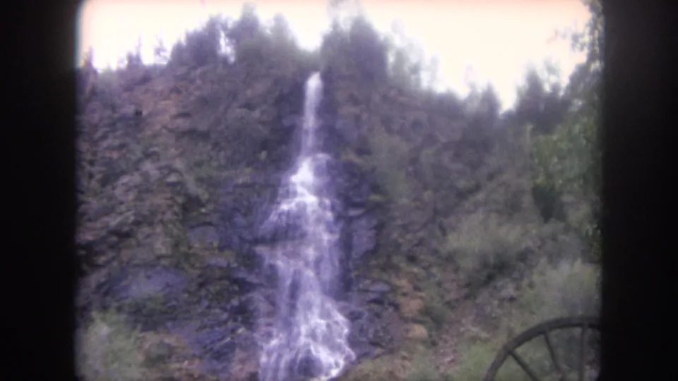 1962 Winter Park home-movie frame with a narrow waterfall descending a rocky, wooded hillside and a partial wooden waterwheel at the edge.