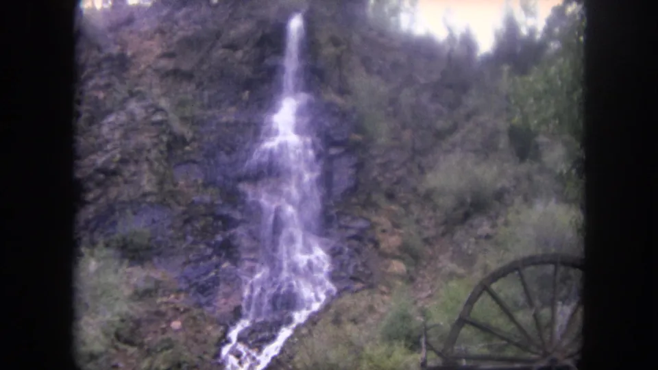 1962 8mm frame from Winter Park, Colorado, showing the waterfall centered on the slope with brush and a partial waterwheel below right.