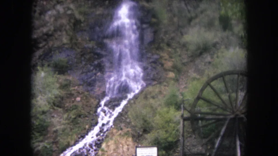 1962 Winter Park, Colorado frame with the waterfall descending toward the lower slope near a large wooden waterwheel and surrounding vegetation.