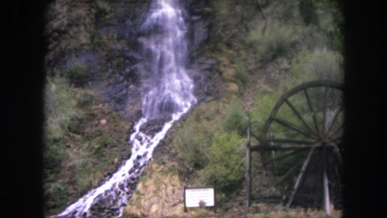 Waterfall running down a rocky hillside beside a wooden waterwheel and small sign.
