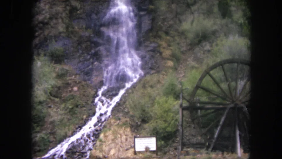 1962 Winter Park, Colorado 8mm frame showing a hillside waterfall, brush, a wooden waterwheel, and a posted sign near the base.