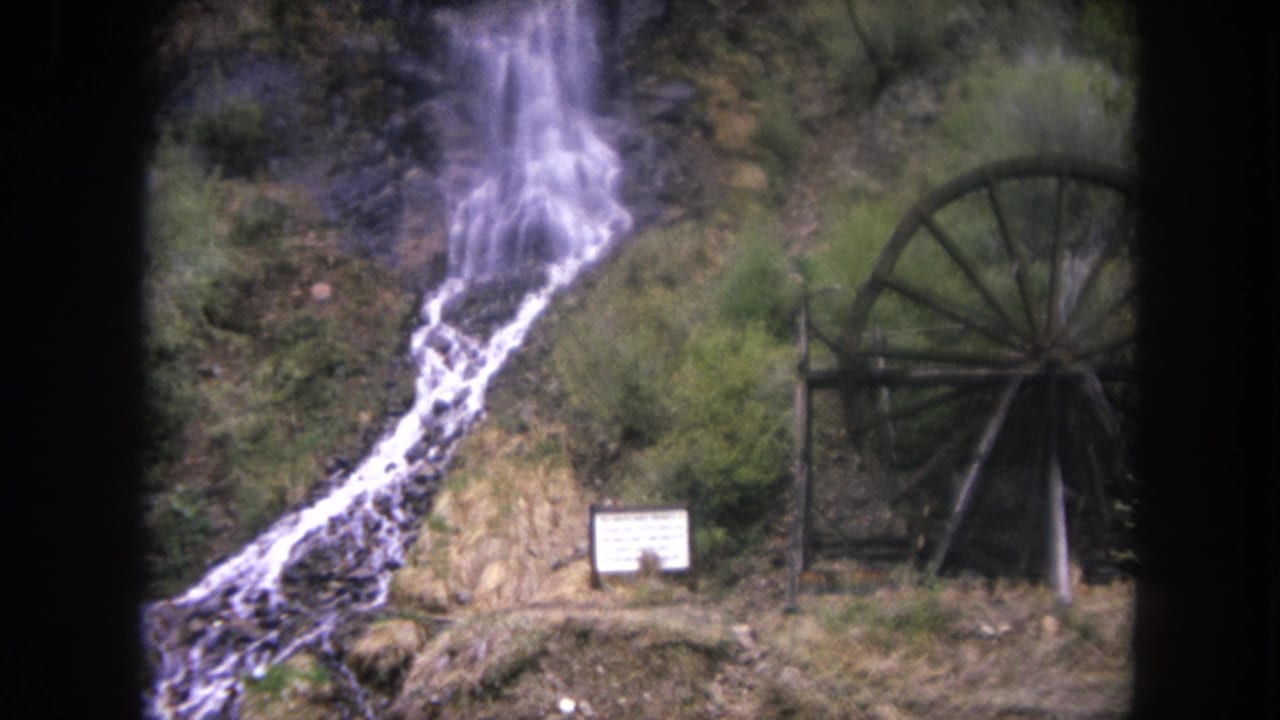 Vintage 8mm view of water cascading down a rocky slope beside a large wooden waterwheel in Winter Park, Colorado.