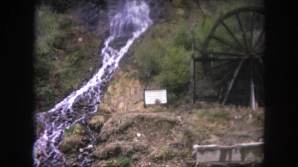 1962 Colorado home-movie frame showing the waterfall flowing toward a stream, with a waterwheel and posted sign in the background.