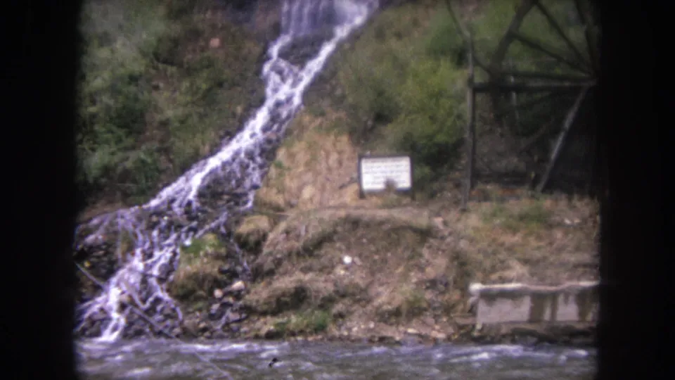 Winter Park, Colorado 8mm frame with stream water in front, a small sign near the bank, and a wooden waterwheel beside the cascade.