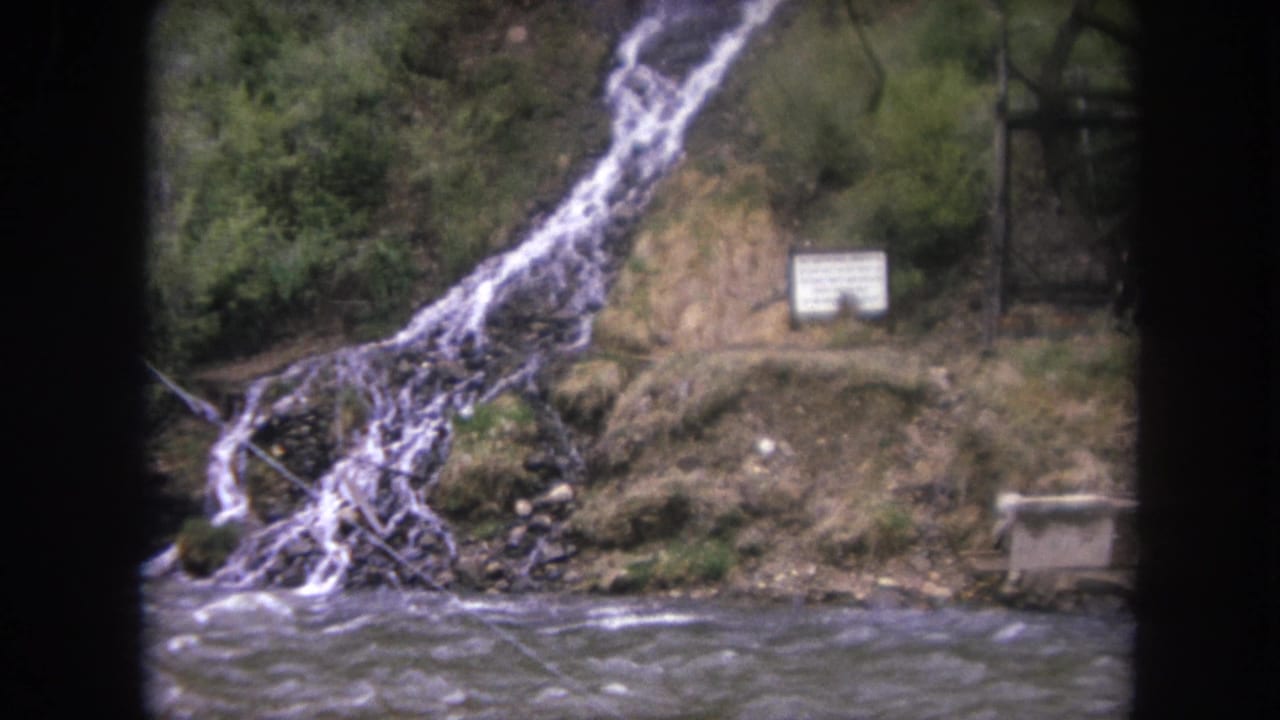 1962 Winter Park, Colorado 8mm frame showing a fast river, small waterfall, rocky bank, trees, and a white sign near the water.