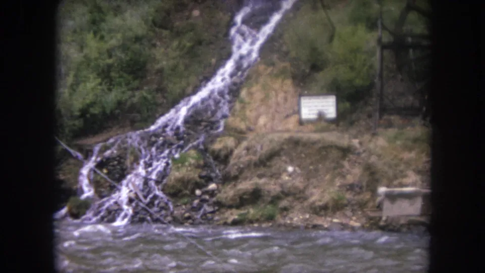 1962 Winter Park, Colorado 8mm frame showing a fast river, small waterfall, rocky bank, trees, and a white sign near the water.