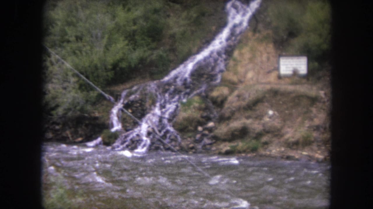 1962 Winter Park, Colorado frame of a small cascade feeding a fast-moving river beside brush, rocks, and a posted white sign.