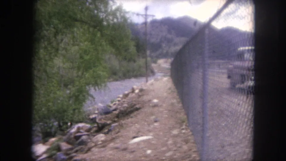1962 Winter Park, Colorado frame showing a chain-link fence along a dirt roadside beside the river, with trees, utility pole, and hills beyond.