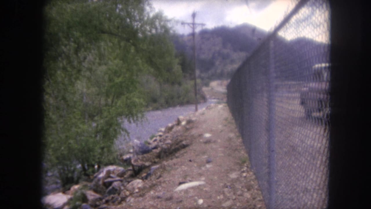 1962 Winter Park, Colorado home-movie frame showing a fence-lined dirt path beside a river and mountain road corridor.