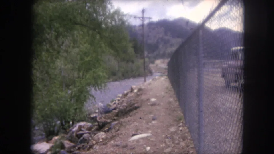 1962 Winter Park, Colorado home-movie frame showing a fence-lined dirt path beside a river and mountain road corridor.